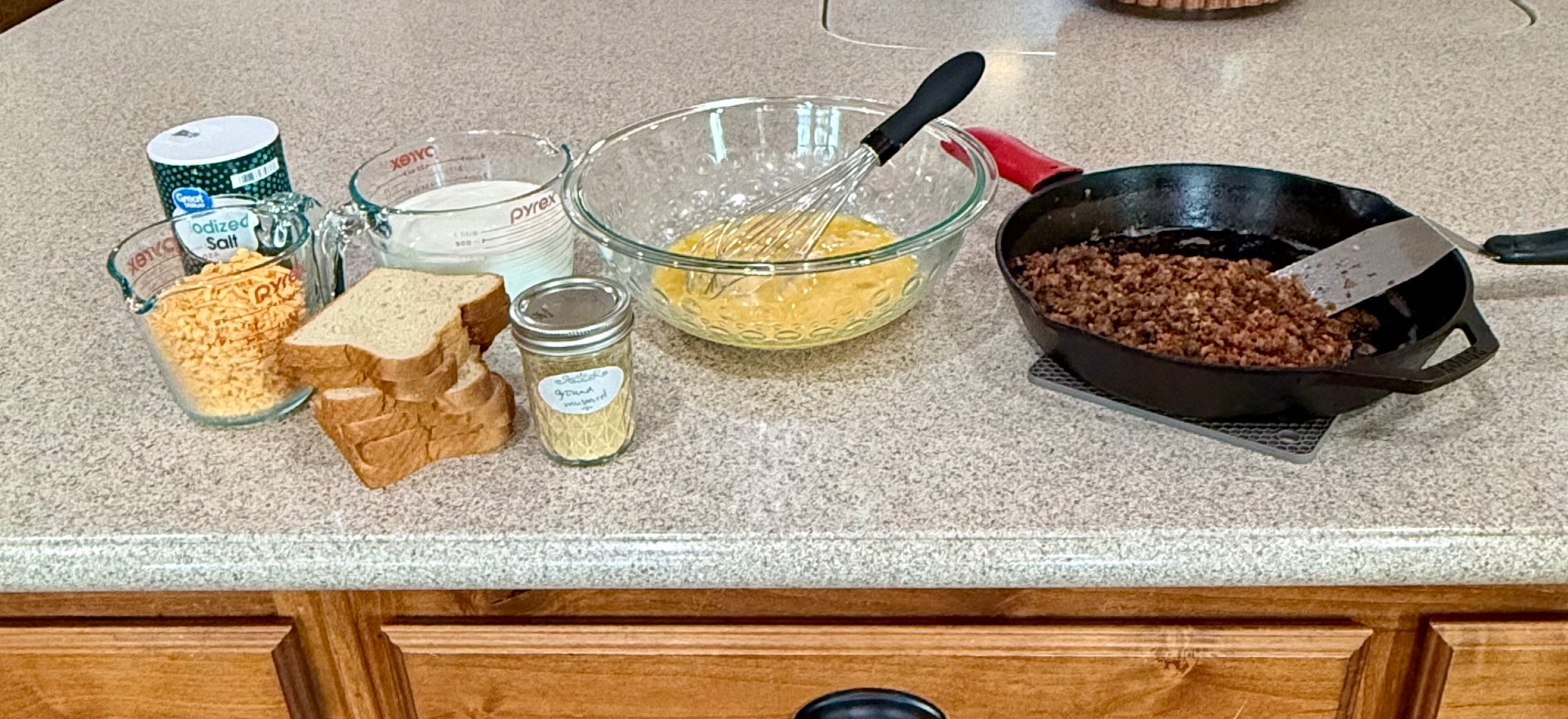 Shredded cheese, salt, milk, sliced bread, a jar of ground mustard, beaten eggs in a glass bowl, and browned sausage in a cast iron skillet set out on the kitchen counter