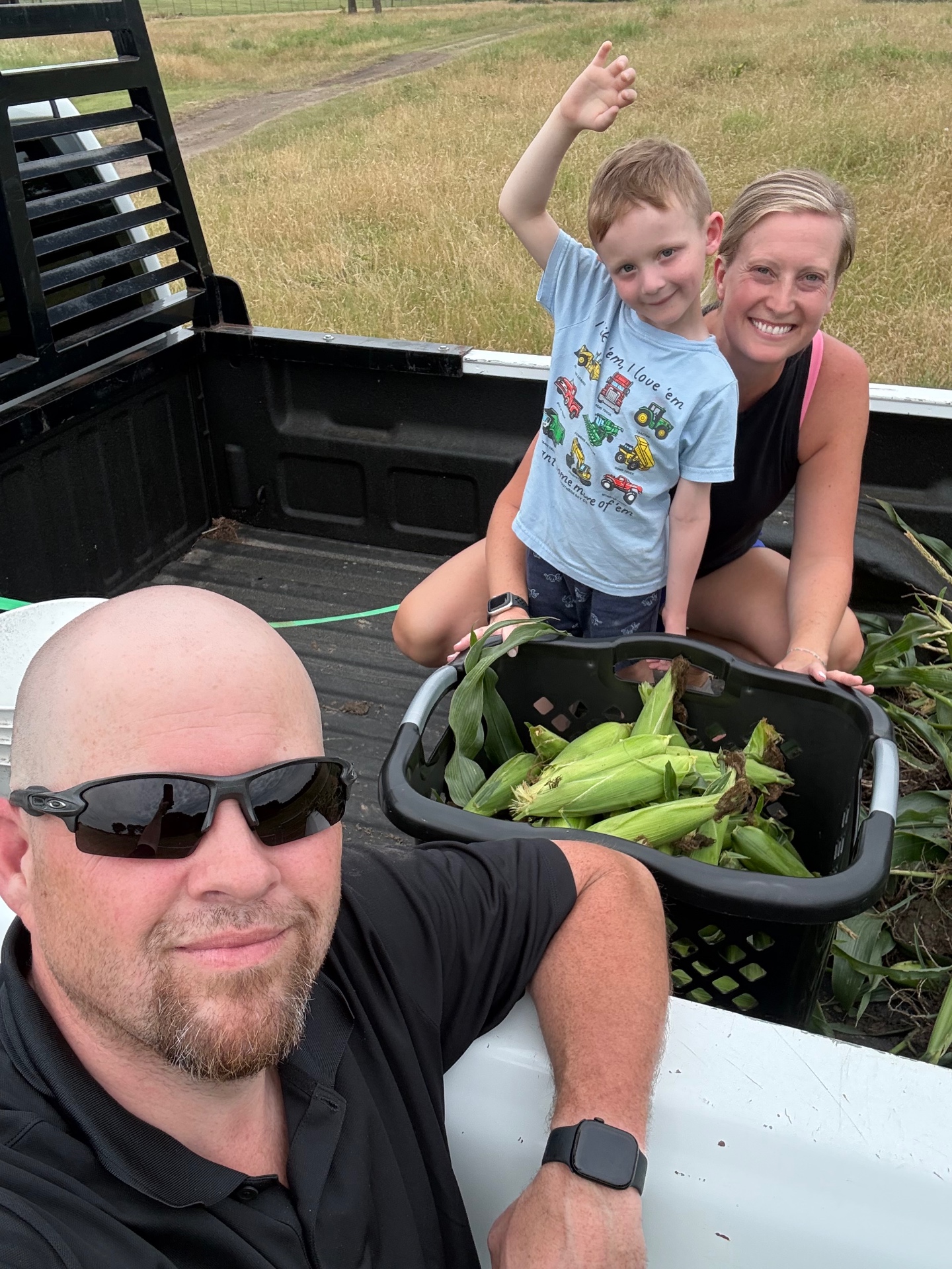 Family in the bed of a truck beside a black basket filled with freshly harvested sweet corn in the husk
