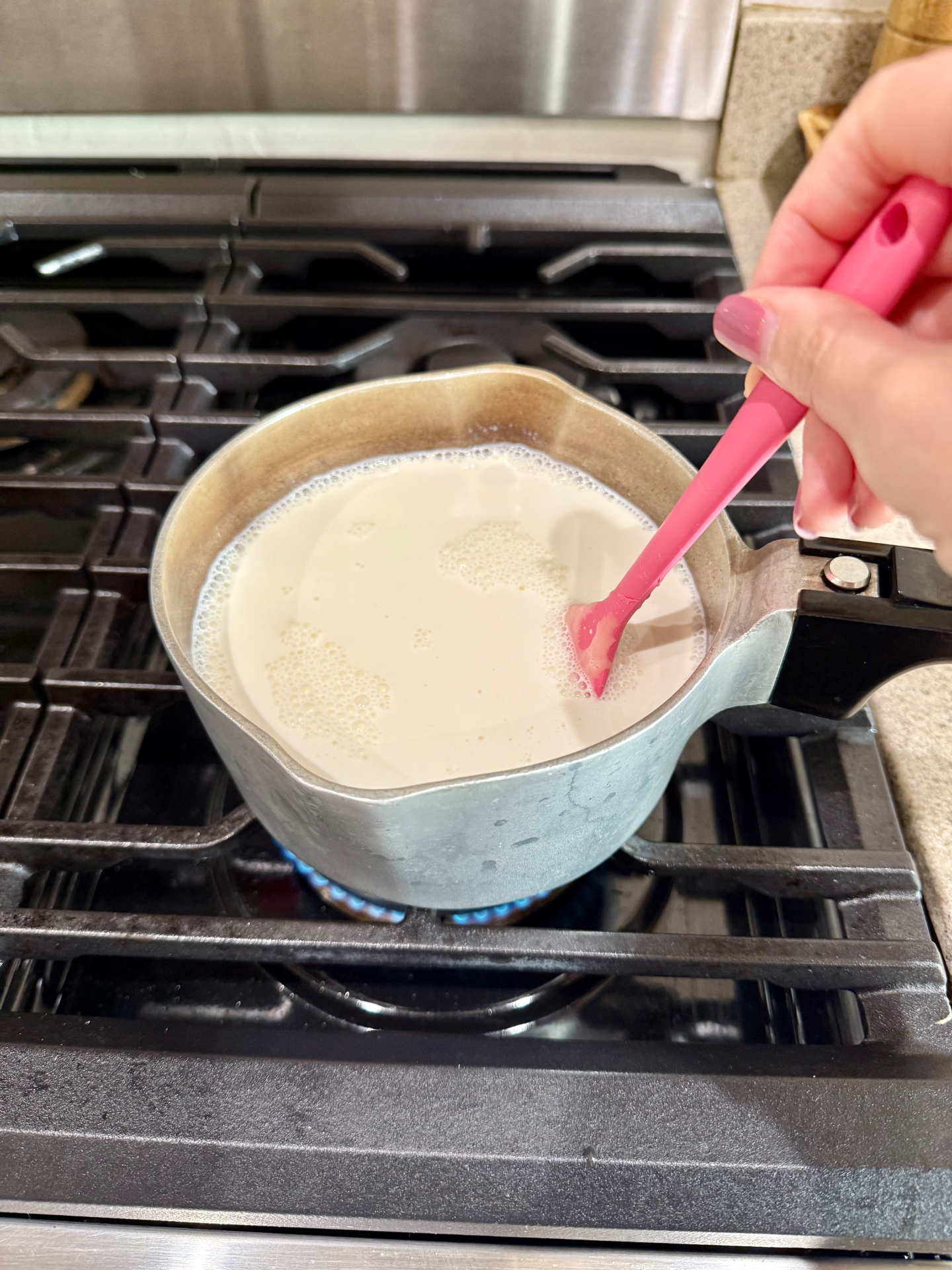 A saucepan of creamer warming on a lit gas stove while it is stirred with a pink silicone spatula.