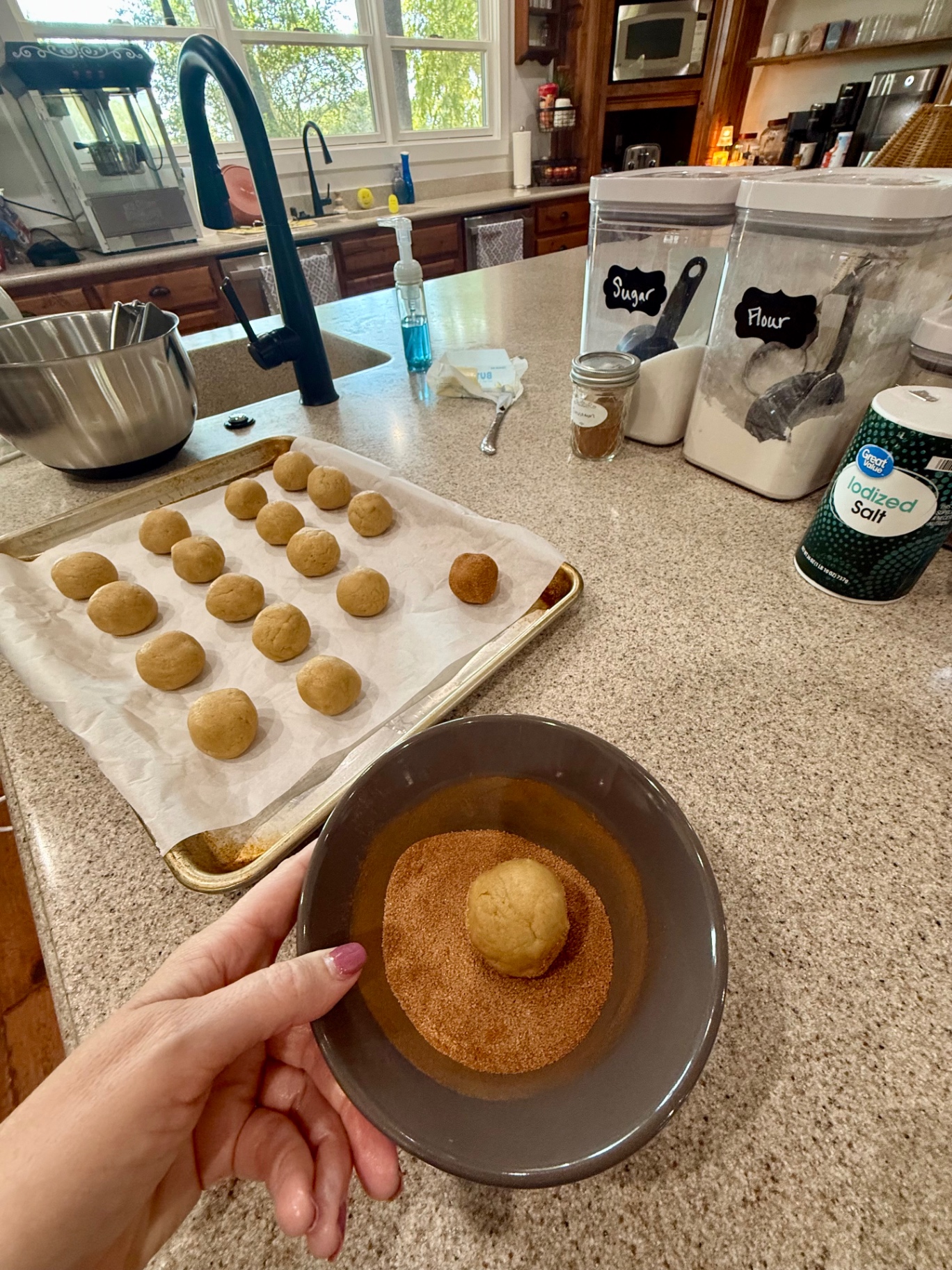 A dough ball being rolled in a bowl of cinnamon sugar, with a tray of scooped dough balls behind it.