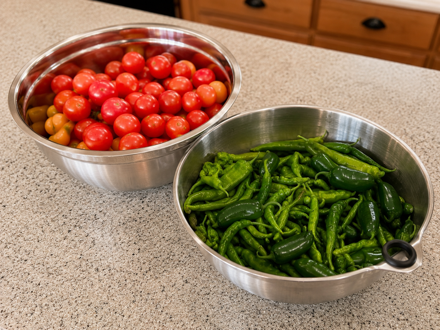 Two stainless steel bowls on a kitchen counter, one full of red cherry tomatoes and one full of green peppers