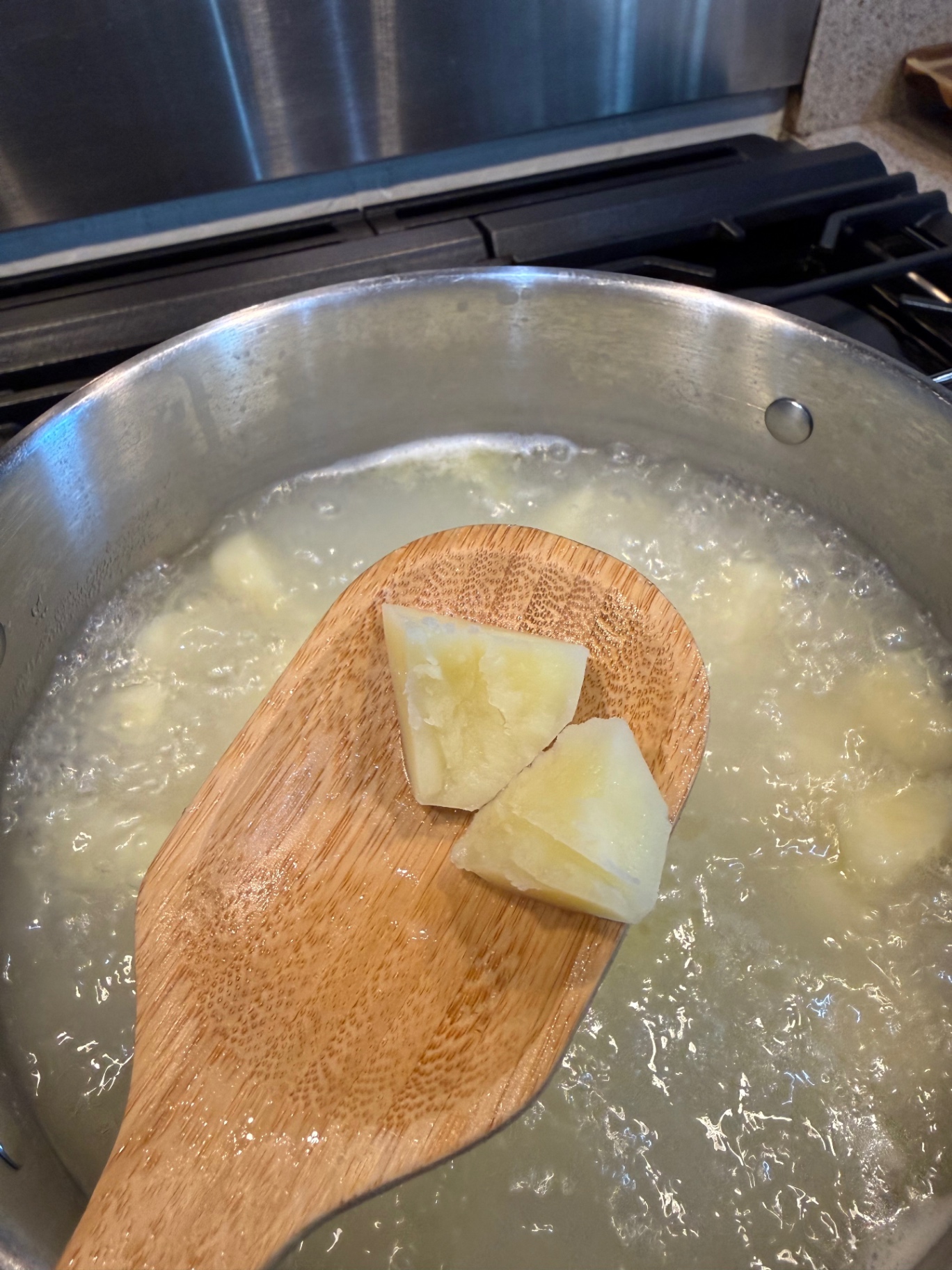 A split, tender potato chunk lifted from the cooking water on a wooden spoon for a doneness check.