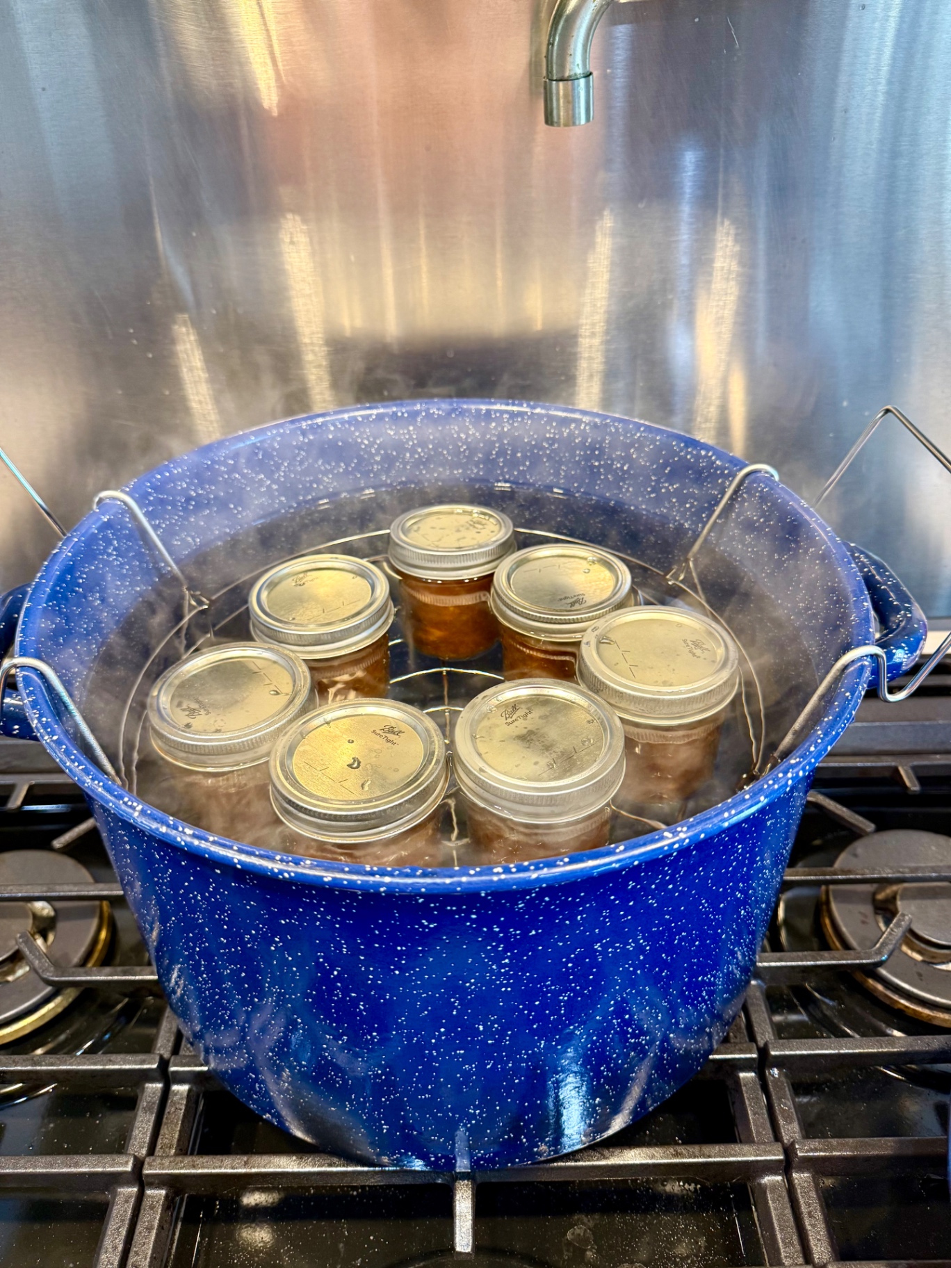 Seven jars of peach topping in a wire rack being lowered into a blue speckled water-bath canner