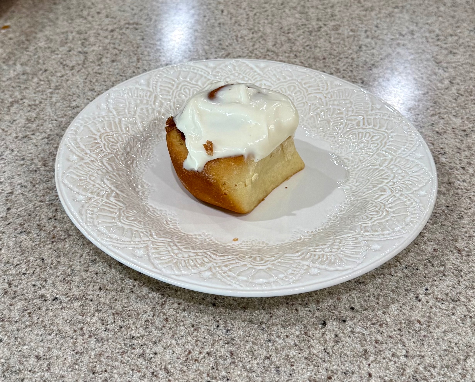 A single frosted cinnamon roll served on a white plate on the kitchen counter