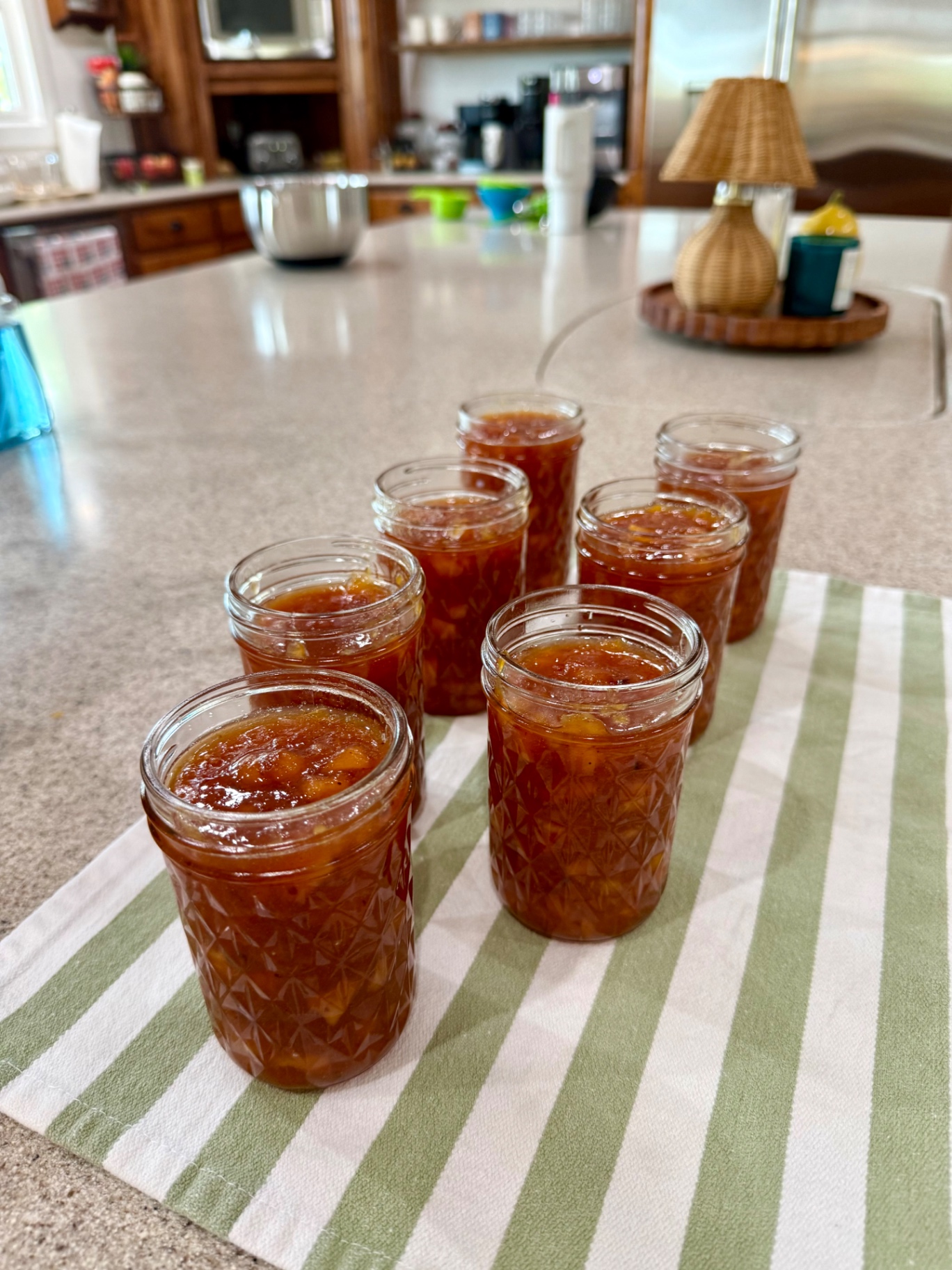 Six hot jars filled with homemade peach topping standing on a striped kitchen towel