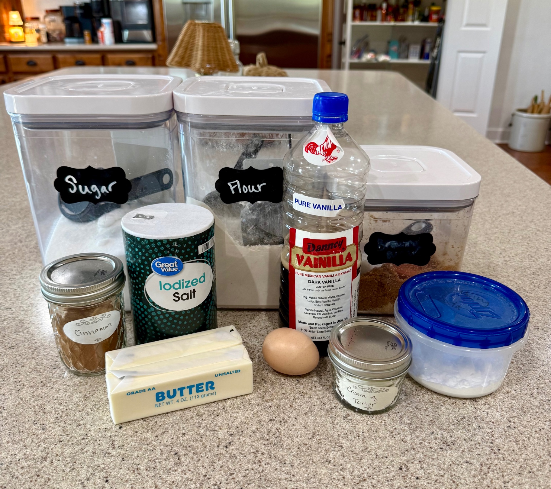 Snickerdoodle ingredients on the kitchen island: sugar and flour canisters, brown sugar, salt, vanilla, cinnamon, butter, an egg, cream of tartar, and baking soda.