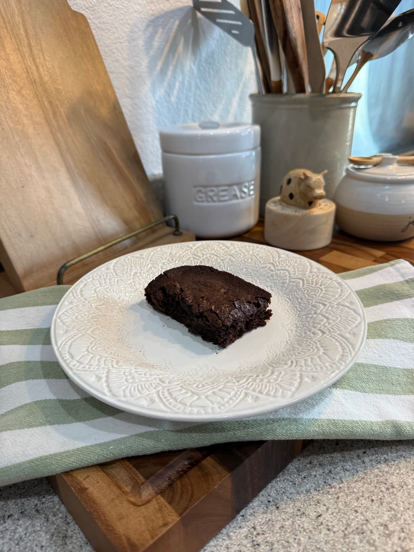A single fudgy brownie on a white embossed plate over a green-and-white striped towel.