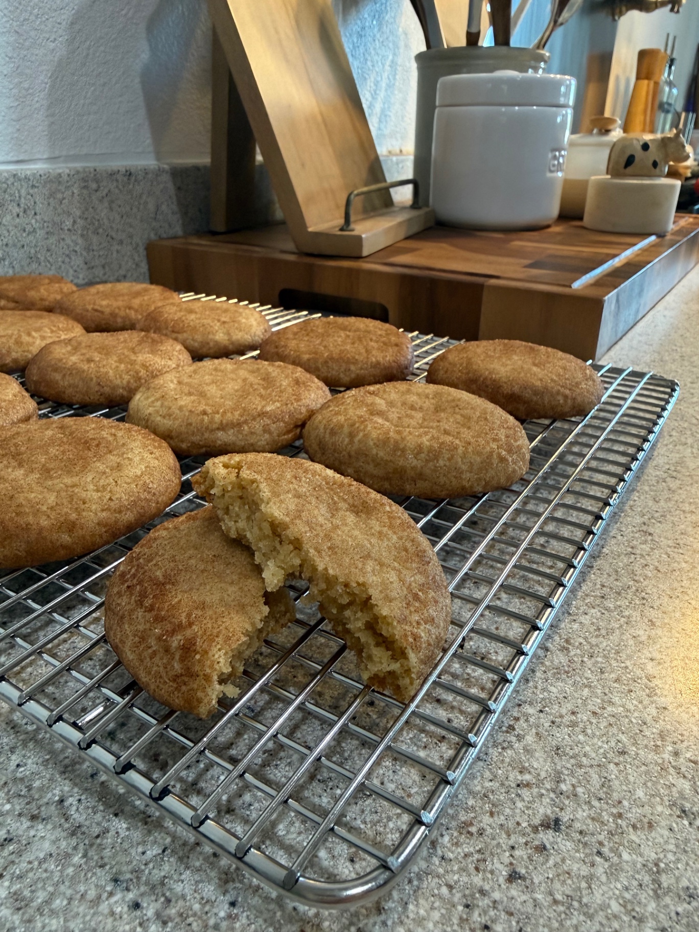 A snickerdoodle broken open on a wire cooling rack to show the soft, chewy center, with more cookies behind it.