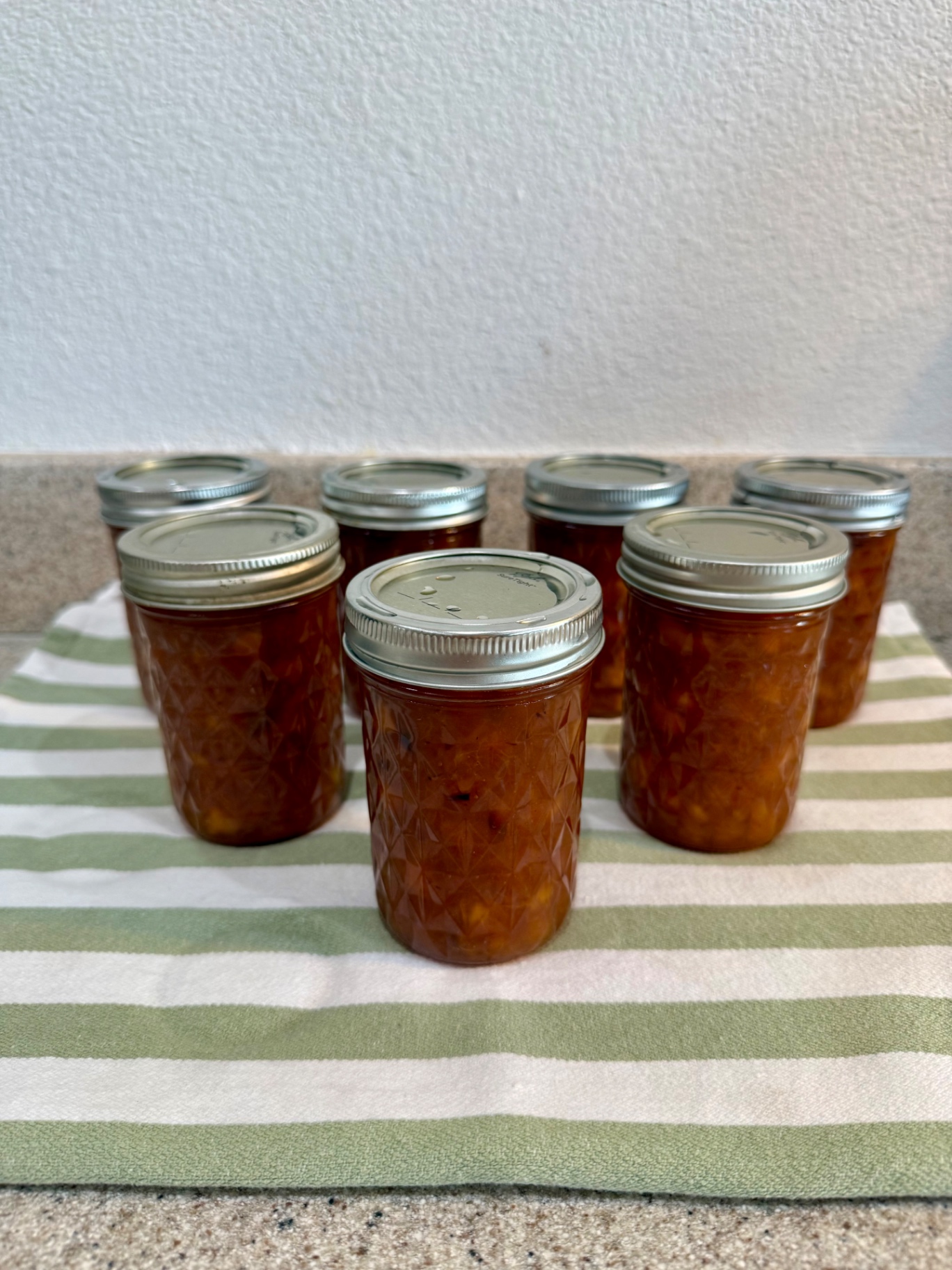 Eight finished jars of homemade peach topping grouped on a green-and-white striped kitchen towel