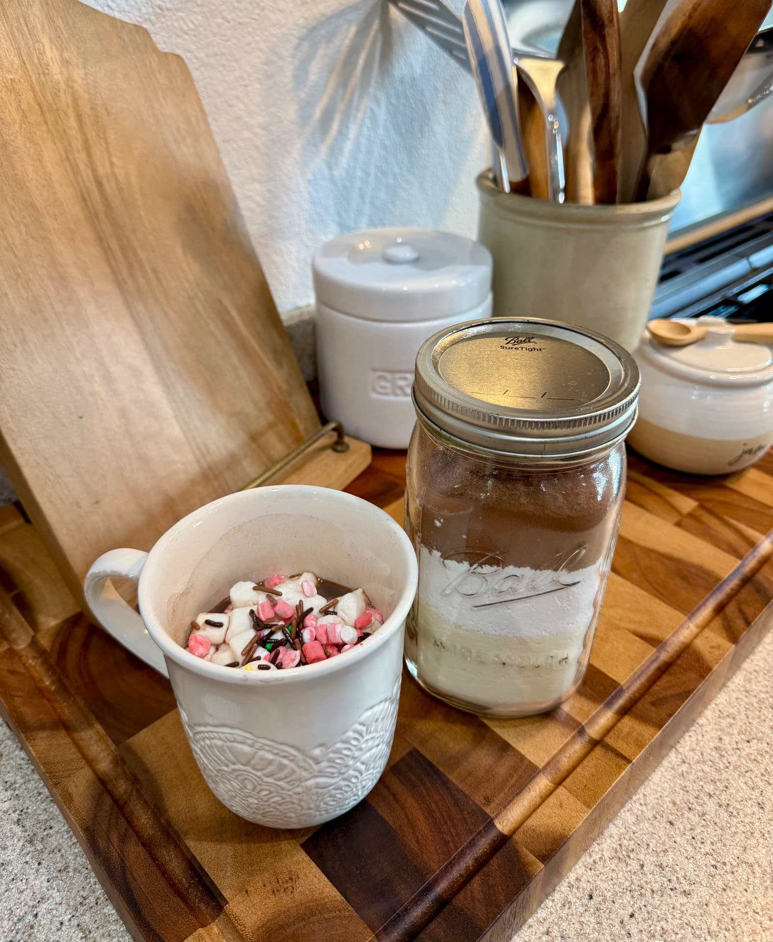 A finished mug of hot cocoa with marshmallows and sprinkles beside a layered quart-size mason jar in the Pasture Threads kitchen.