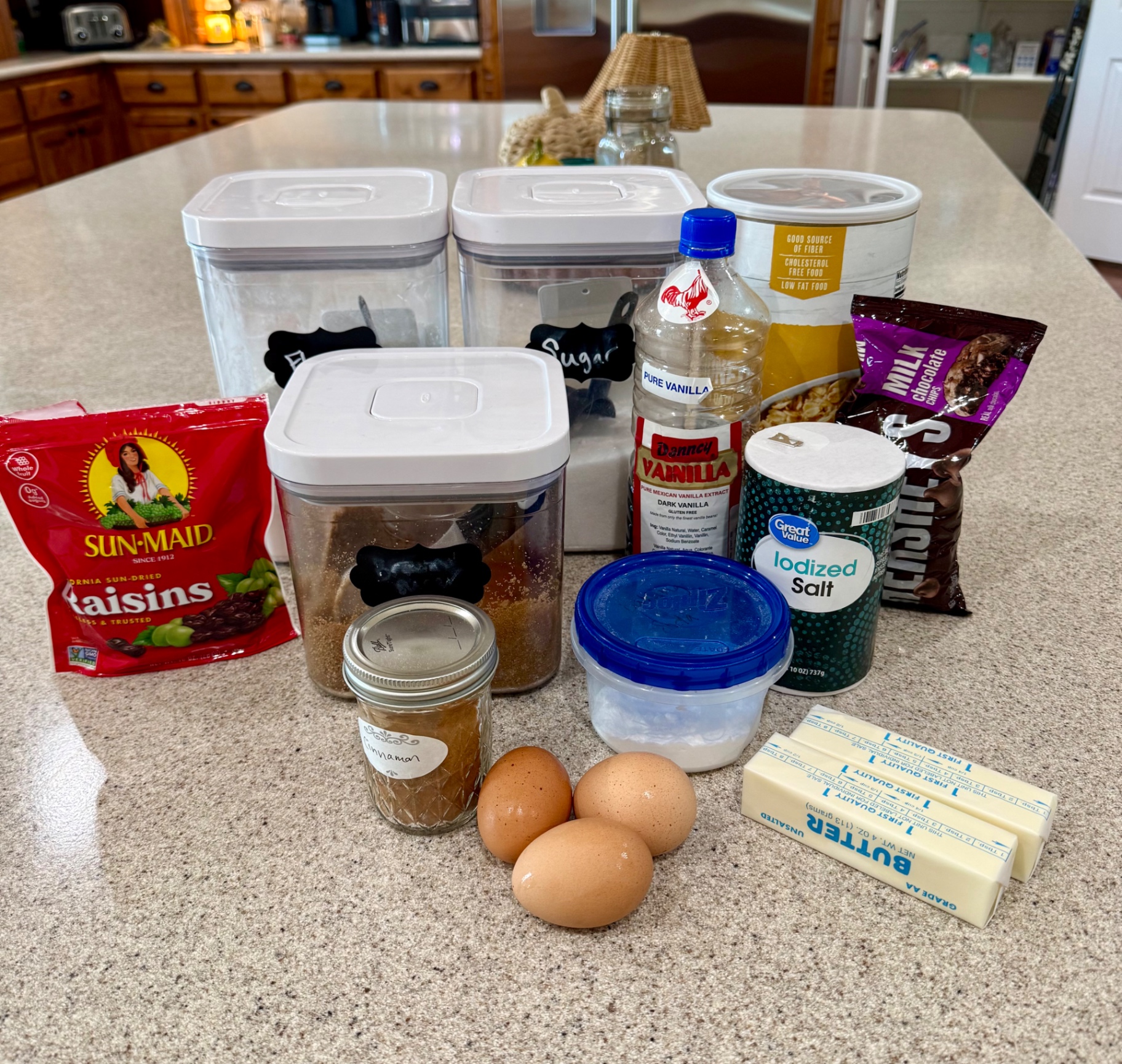 Oatmeal cookie ingredients gathered on the kitchen counter, including butter, eggs, oats, flour, sugars, vanilla, cinnamon, salt, raisins, and chocolate chips.