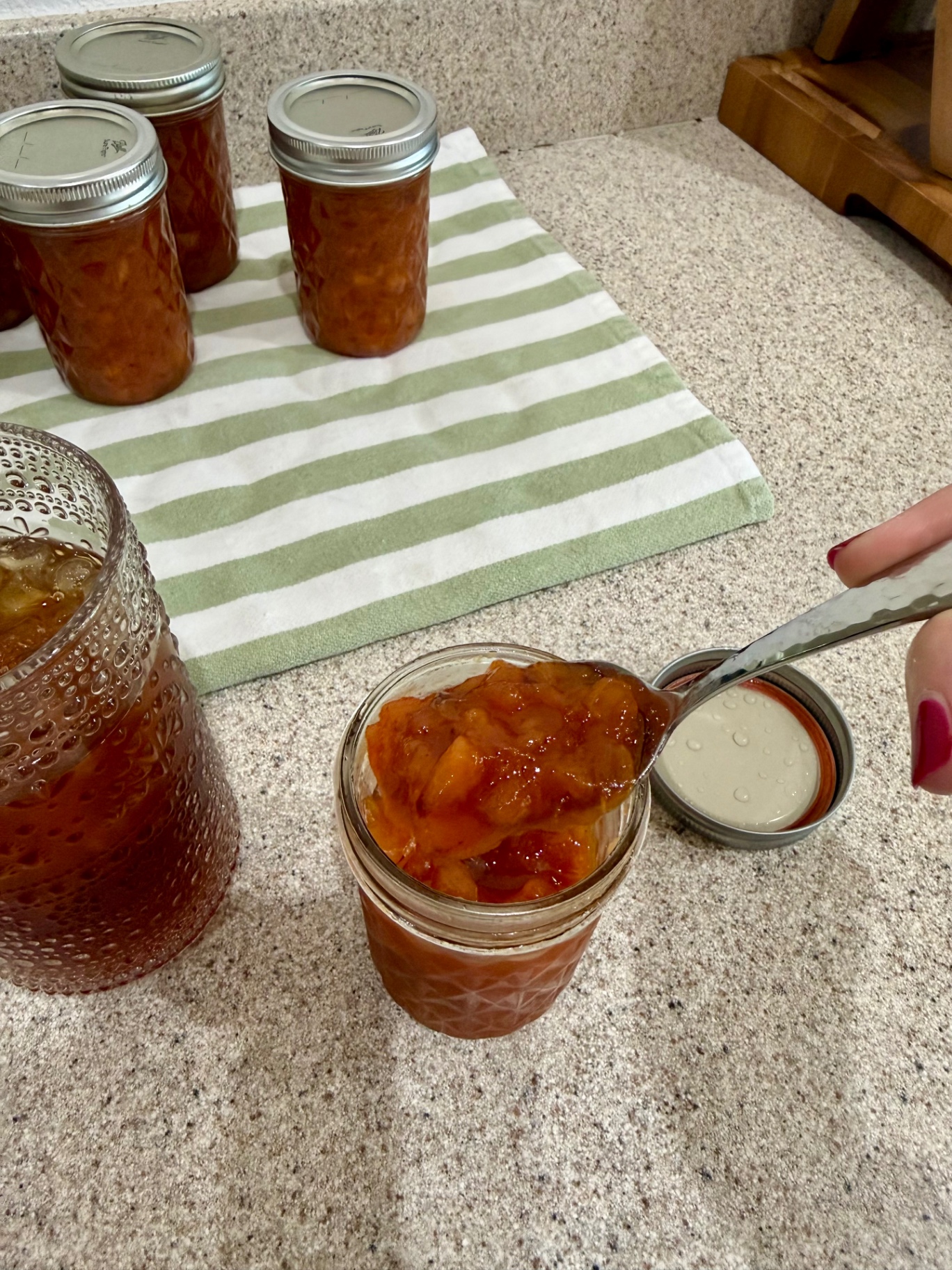 A spoonful of soft peach topping held over an open jar beside a glass of iced tea