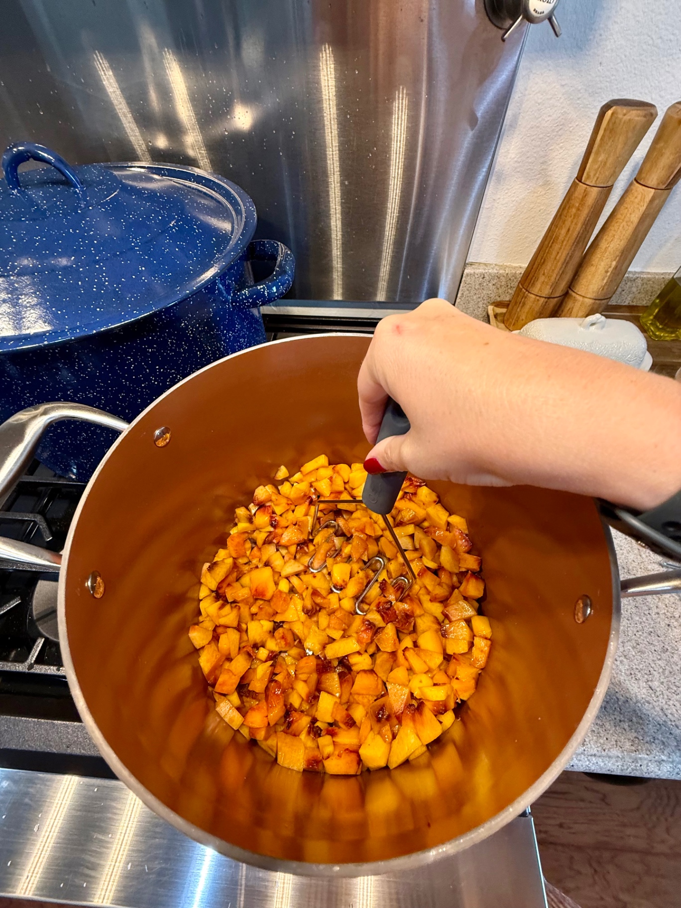 Diced yellow peaches being gently mashed with a potato masher in a large copper-colored pot