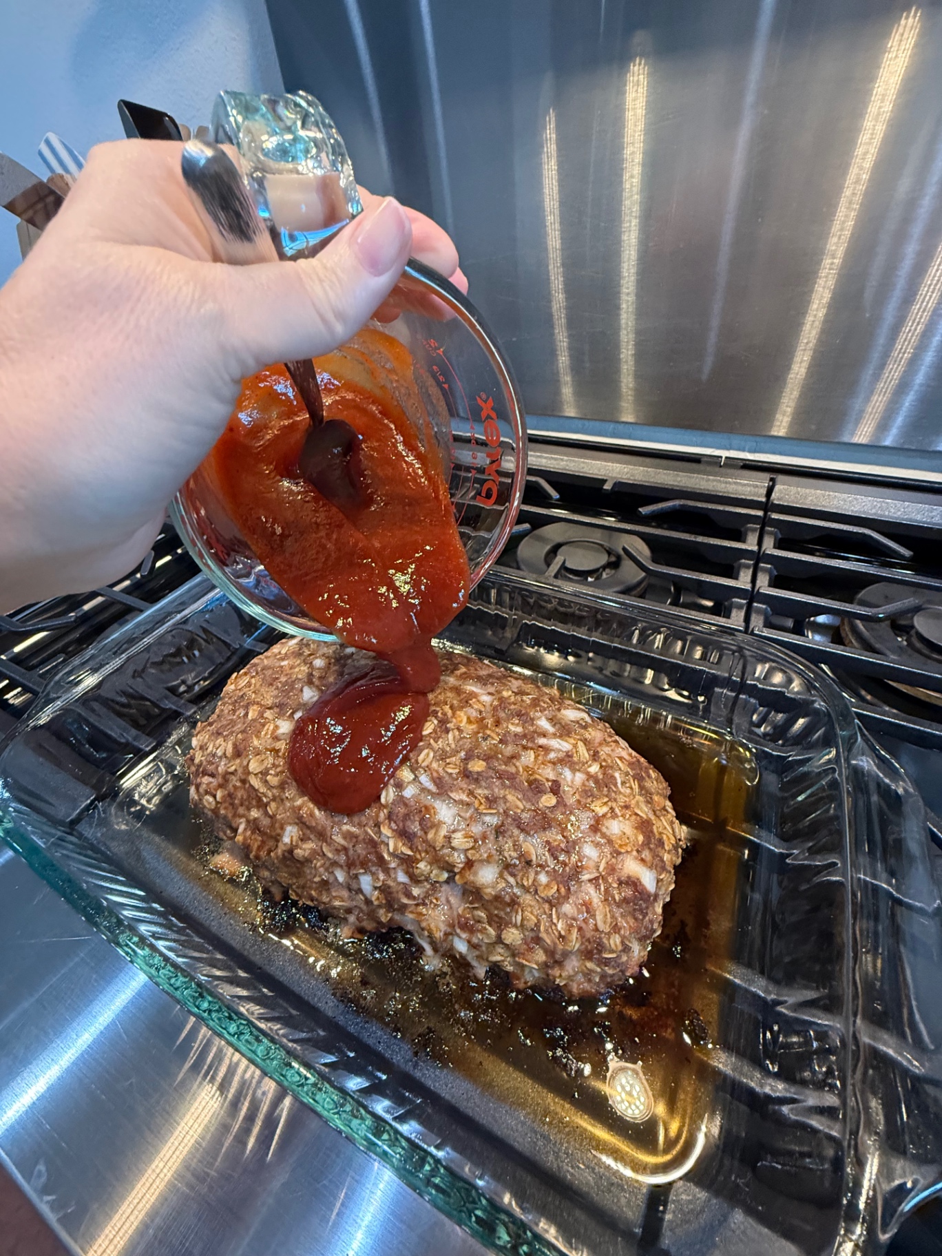 Pouring tomato glaze from a glass measuring cup over the meatloaf in its baking dish.