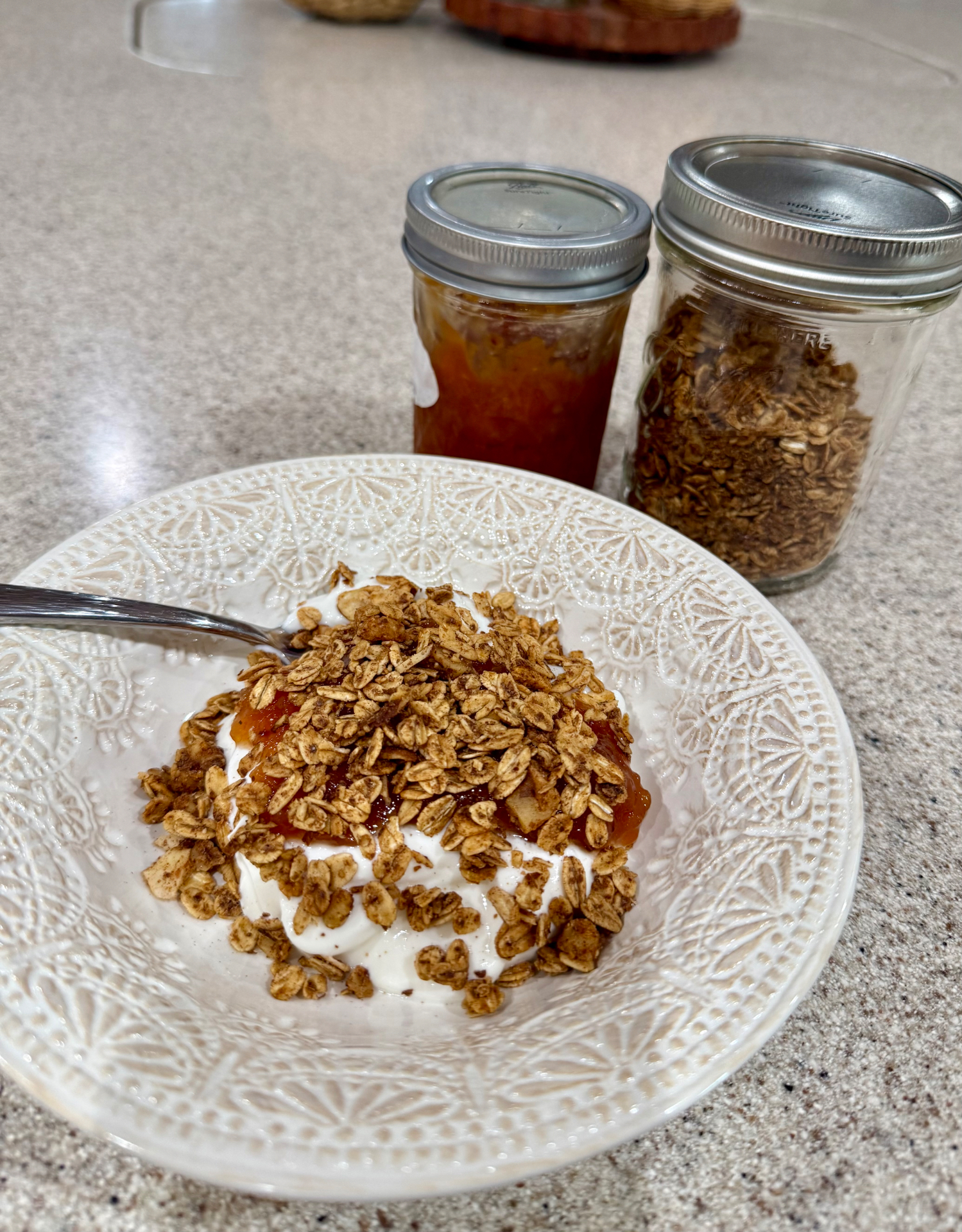 Honey cinnamon walnut granola over Greek yogurt with homemade peach topping, with jars of granola and peach topping behind the bowl.