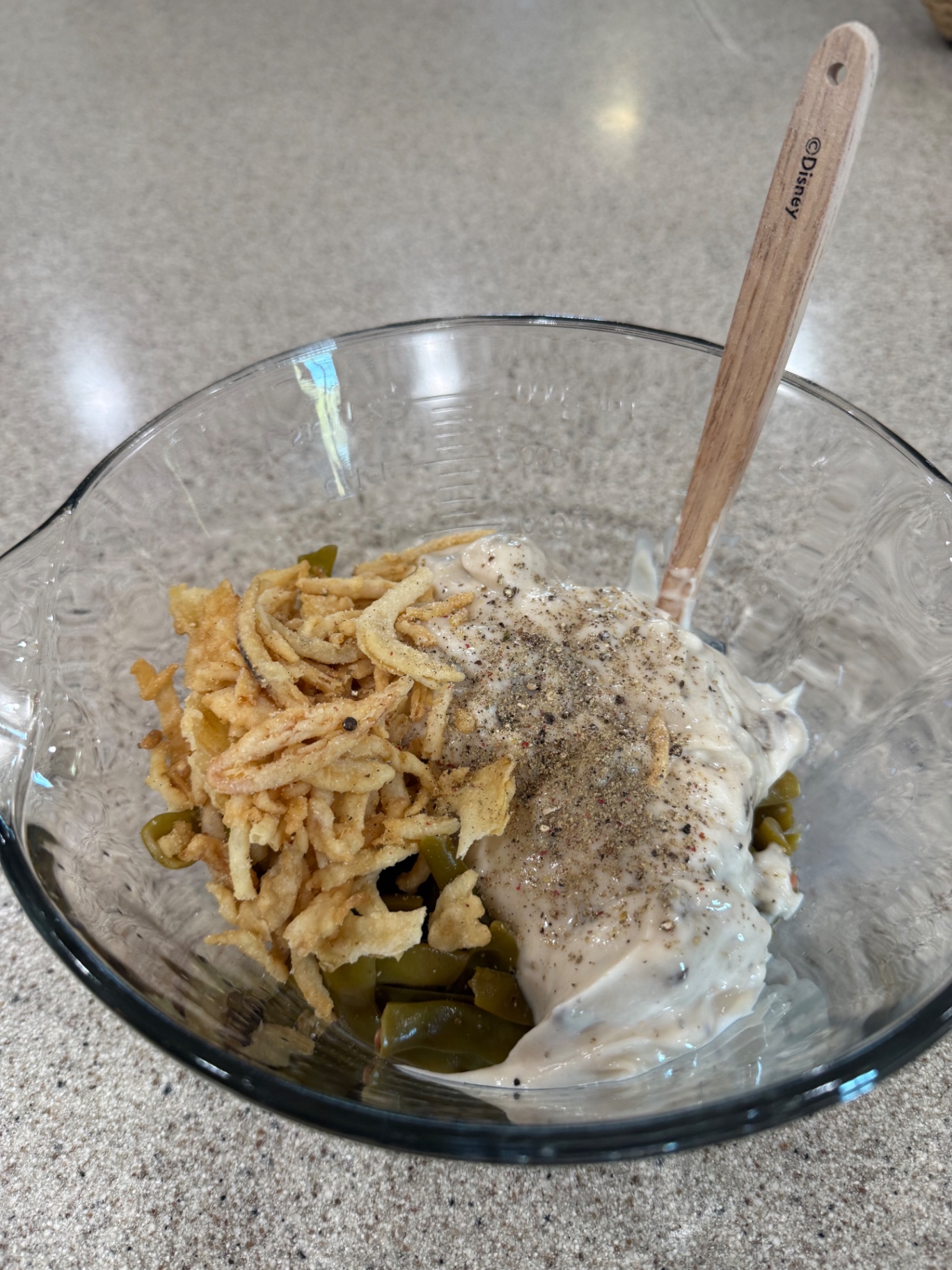 Green beans, cream of mushroom soup, seasoning, and crispy fried onions in a glass bowl before stirring.
