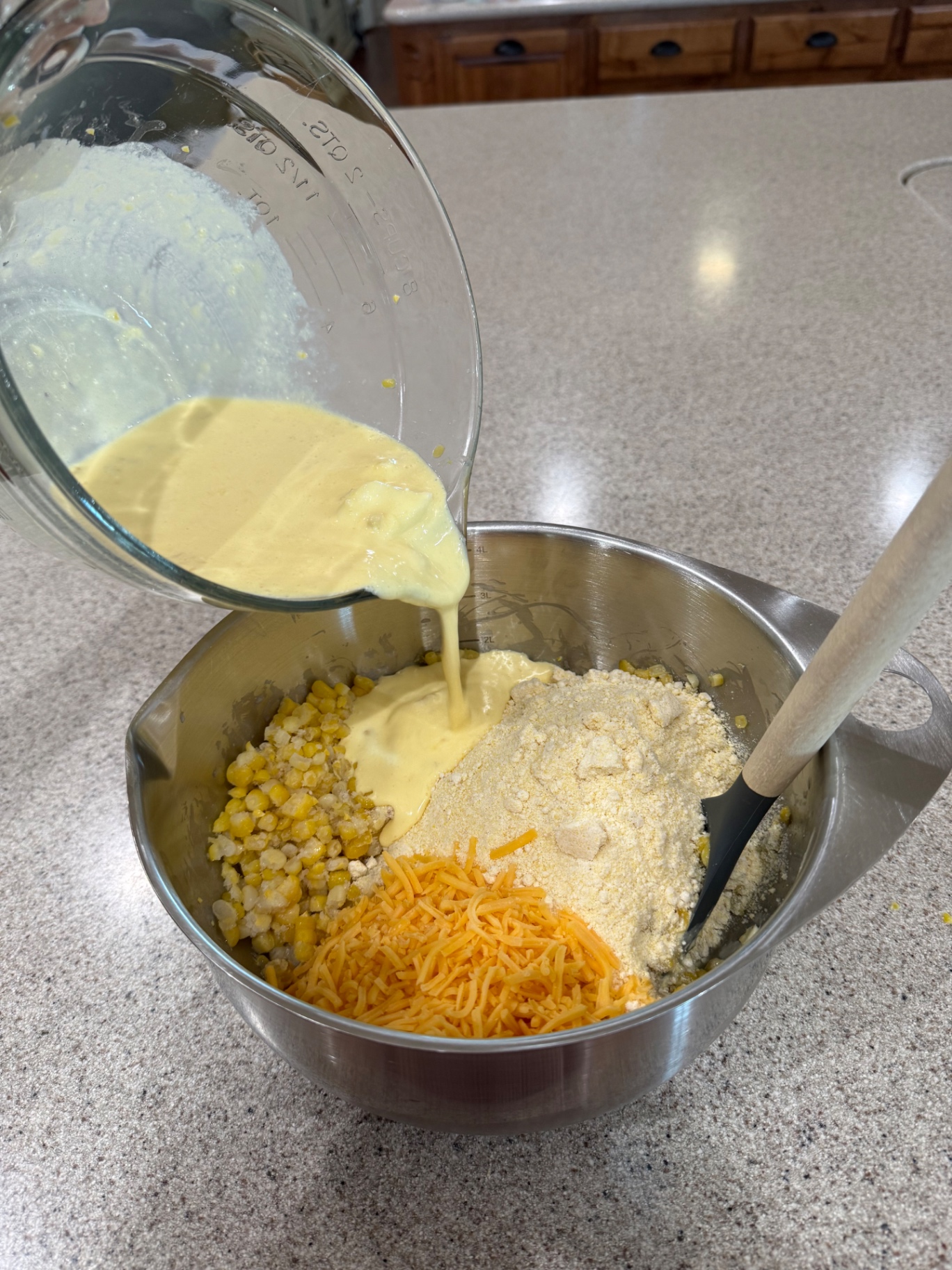 Whisked wet ingredients being poured into corn, Jiffy Corn Muffin Mix, and cheddar in a metal bowl.